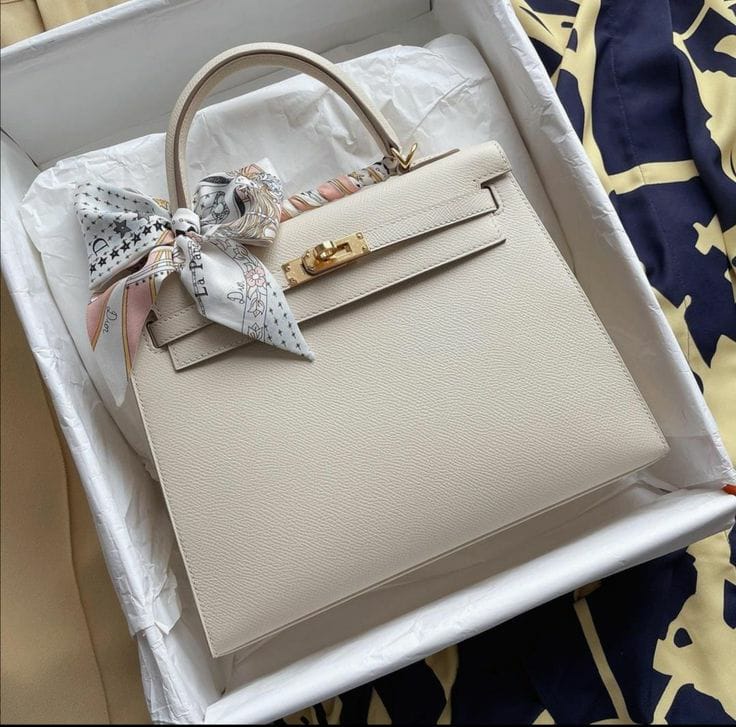 Hermès Kelly Sellier in a neutral shade like Craie or Nata with gold-tone hardware, accented with a tied twilly scarf.